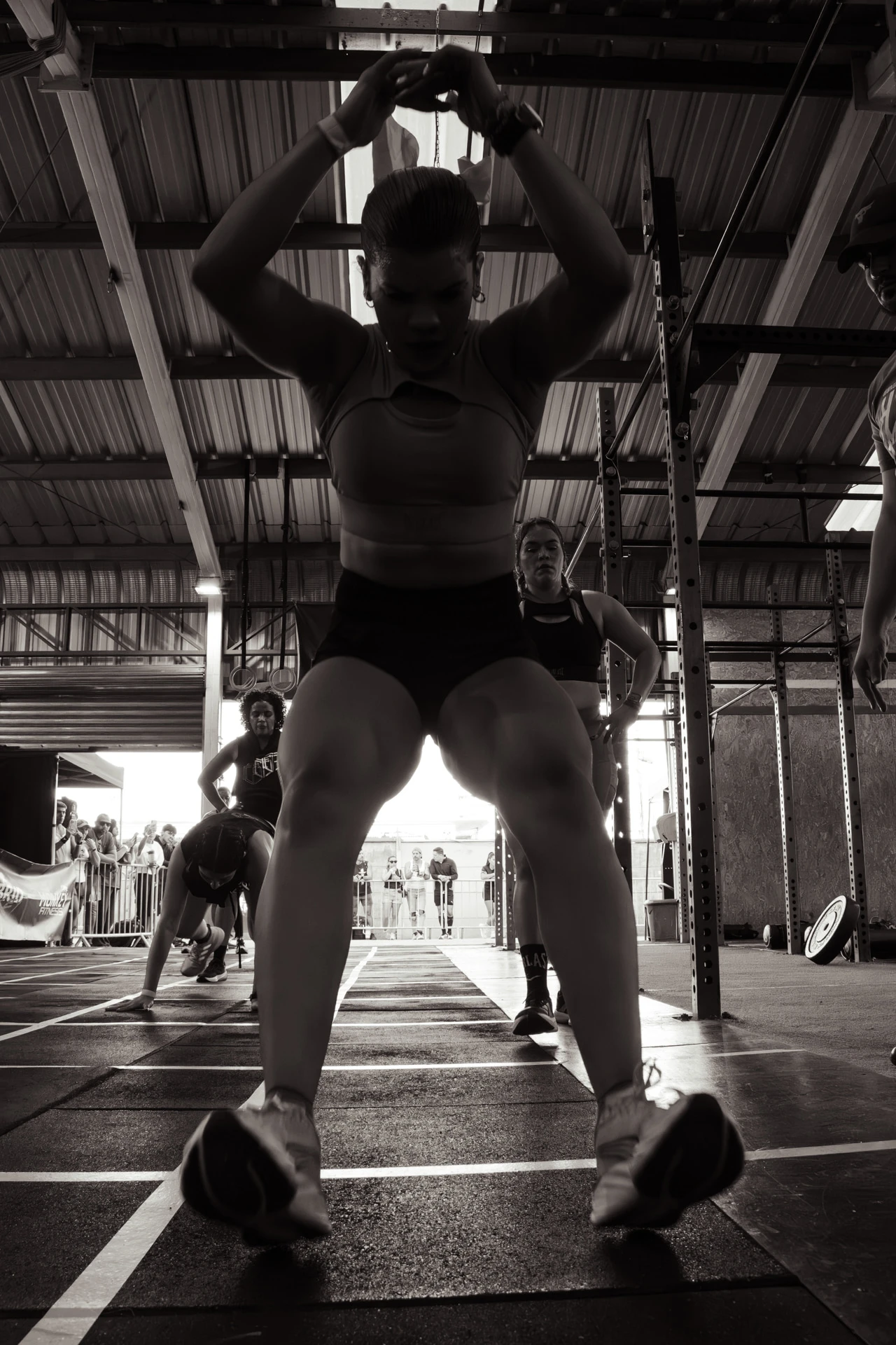 Captada desde un ángulo bajo y en blanco y negro, una atleta aterriza tras un burpee broad jump durante Hybrid Monkey V4, con otros participantes al fondo.