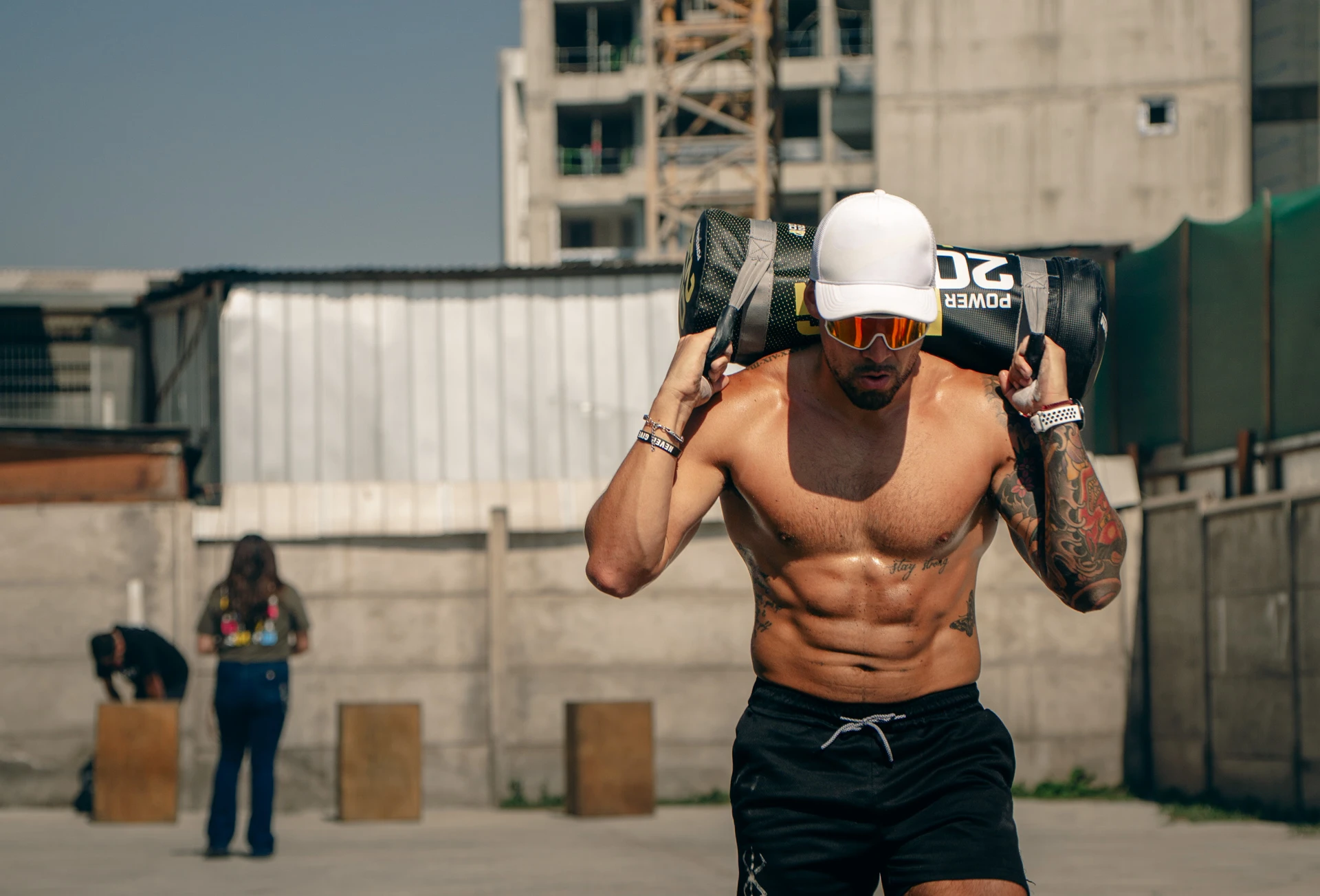 Con gorra blanca y gafas de sol, un atleta lleva un saco de 20 kg sobre los hombros mientras realiza la estación de estocadas de Hybrid Monkey V2.